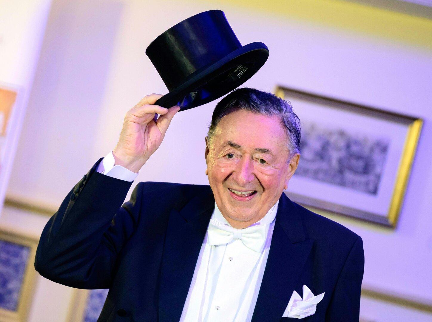 Ein lächelnder Mann im Smoking mit Fliege hält einen Zylinderhut hoch.