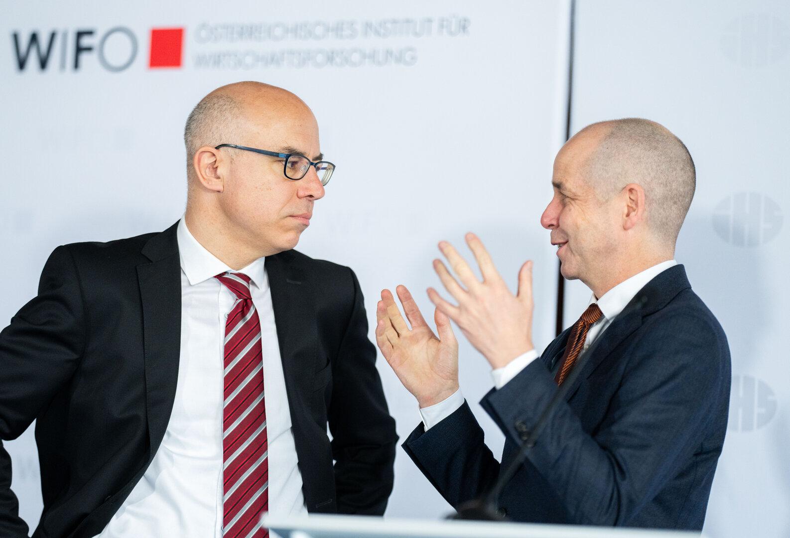 Zwei Männer in Anzügen unterhalten sich vor dem Logo des WIFO.