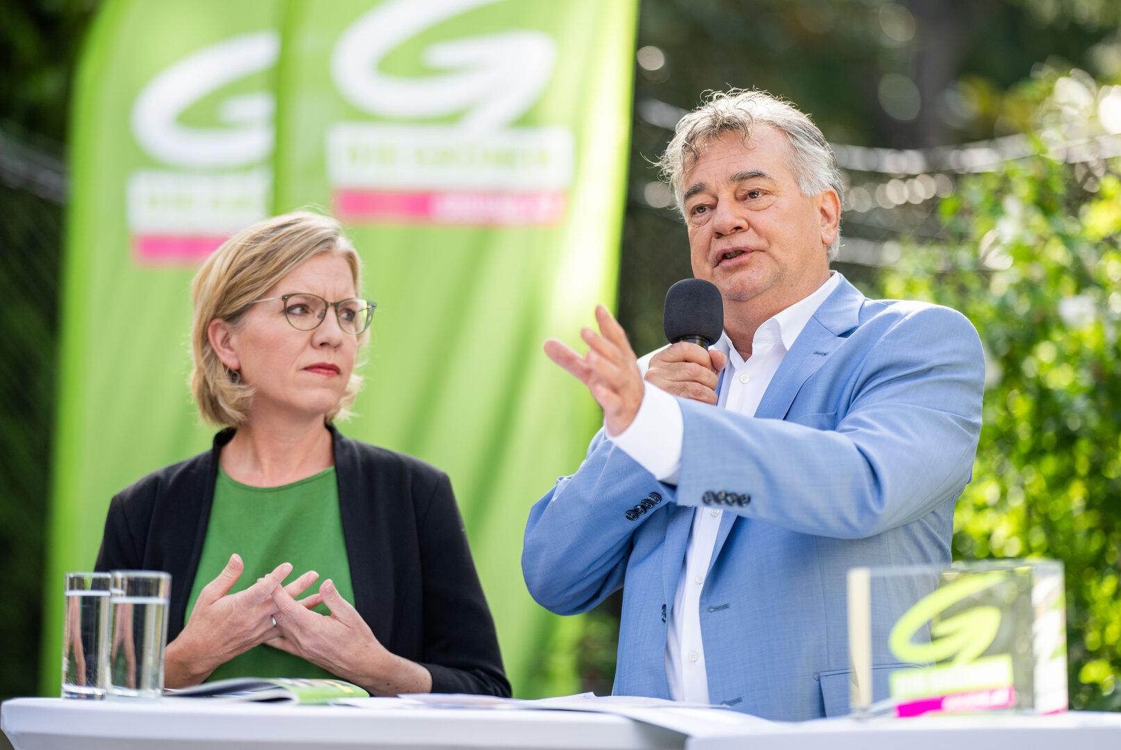 Ein Mann im blauen Anzug spricht am Mikrofon, während eine Frau aufmerksam zuhört.