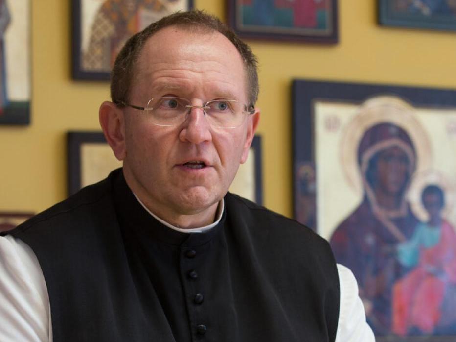 Ein Mann mit Brille und Klerikerhemd vor einer Wand mit Bildern.