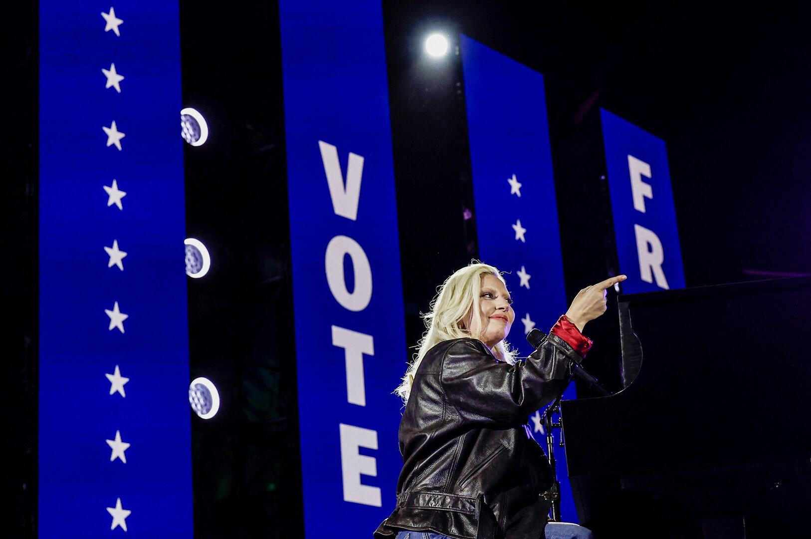 Lady Gaga sitzt am Klavier vor Bannern, die zum Wählen auffordern.