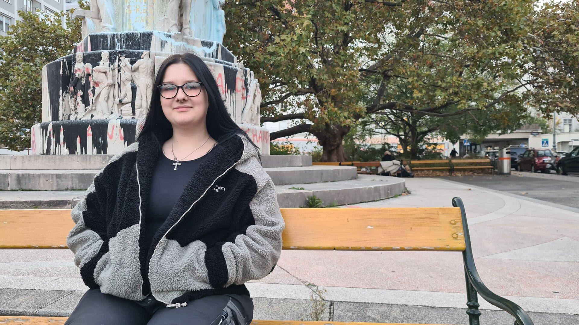 Eine junge Frau mit Brille sitzt auf einer Bank vor einem Denkmal.