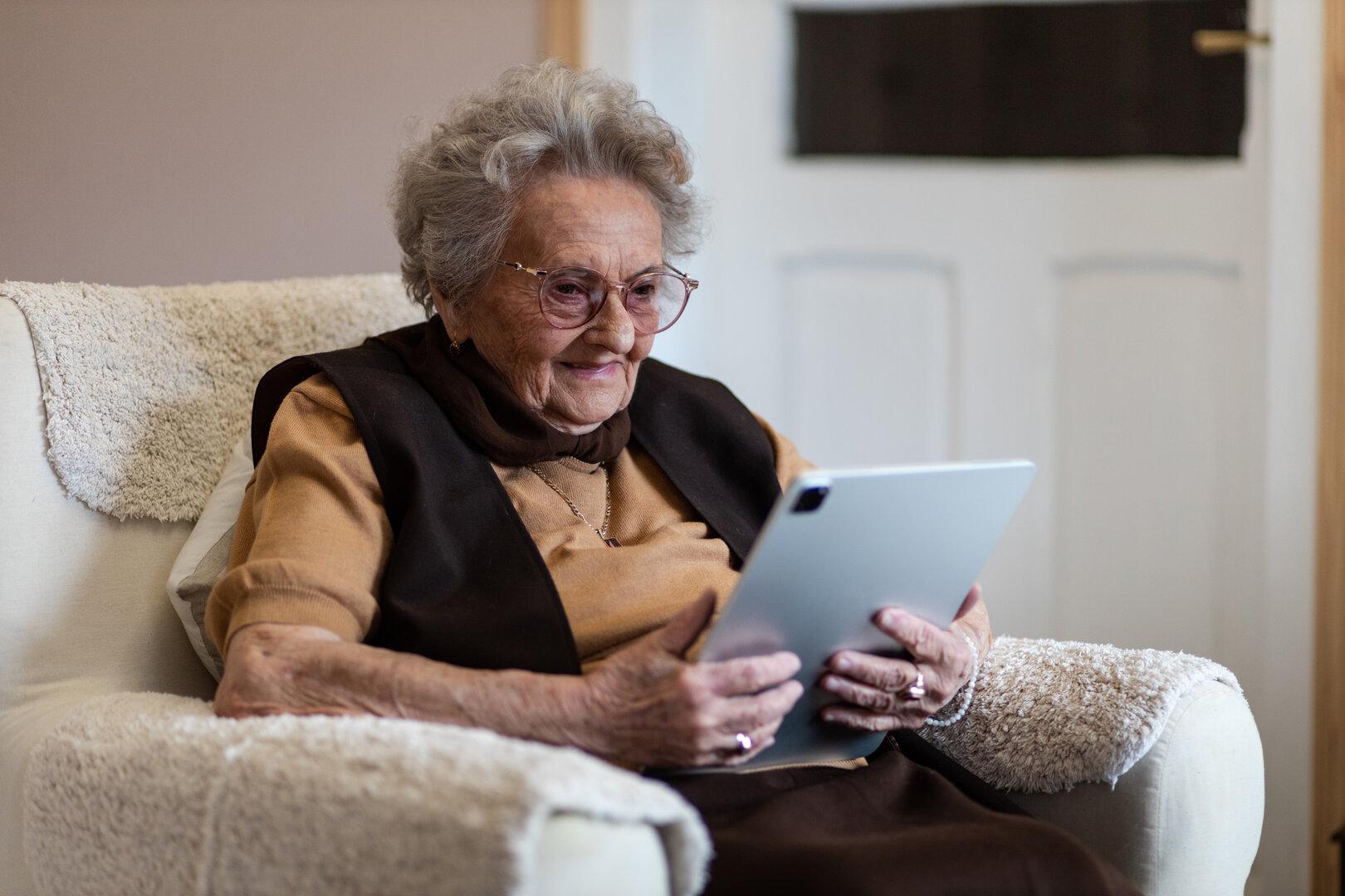Eine ältere Frau sitzt in einem Sessel und benutzt ein Tablet.