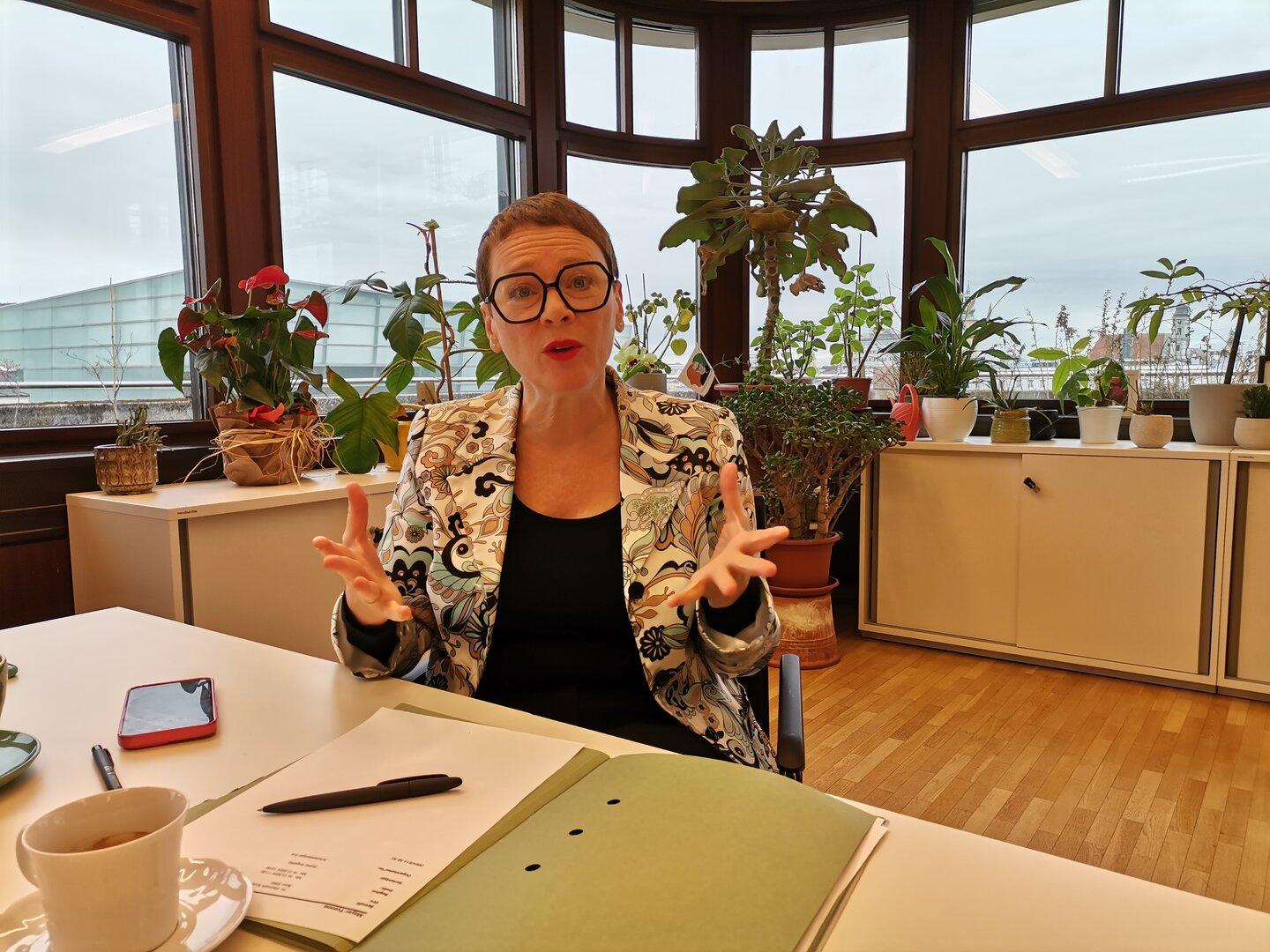 Eine Frau mit Brille gestikuliert an einem Schreibtisch voller Pflanzen.