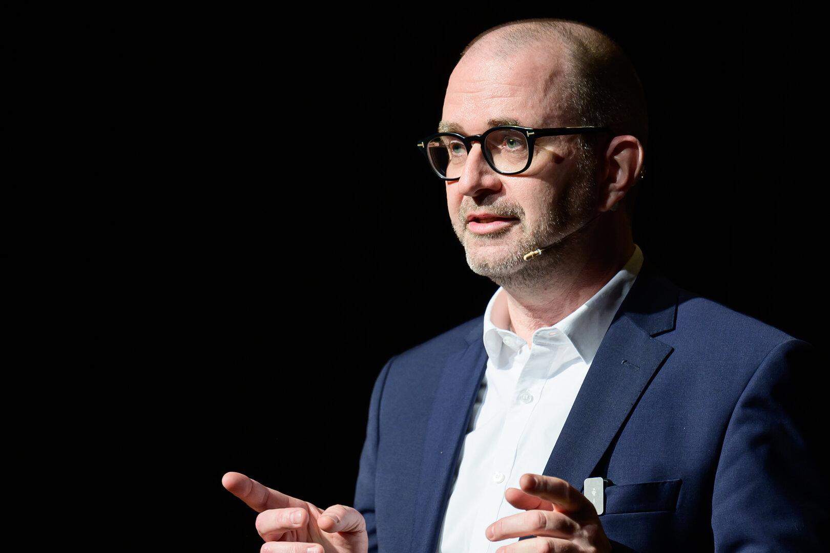 Ein Mann mit Brille und Anzug gestikuliert vor einem dunklen Hintergrund.