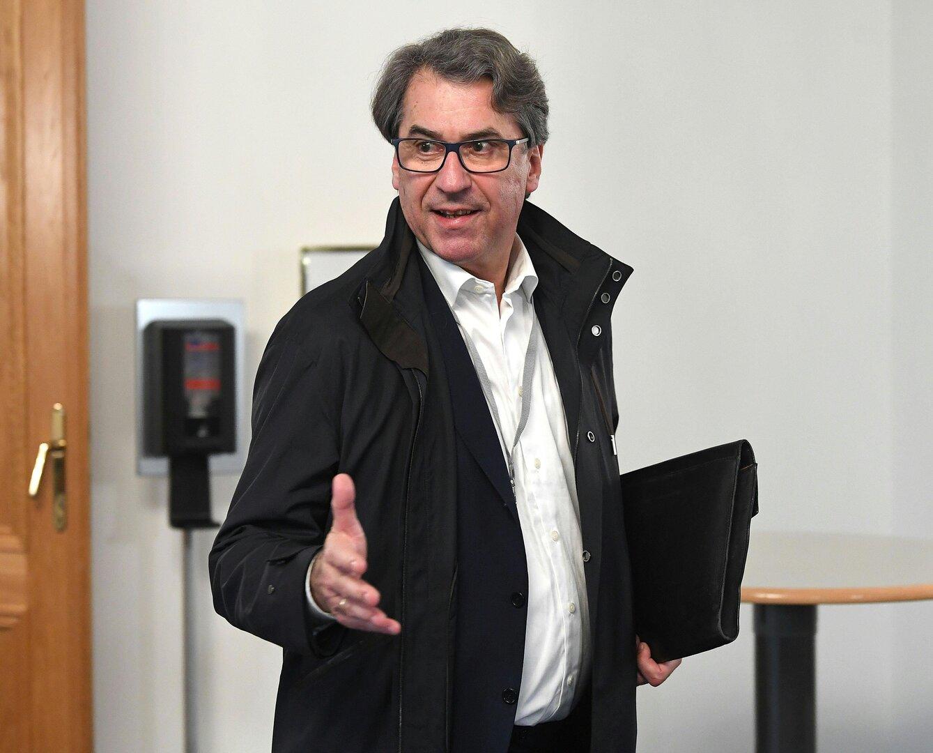 Ein Mann mit Brille und Jacke gestikuliert und hält eine schwarze Aktentasche.