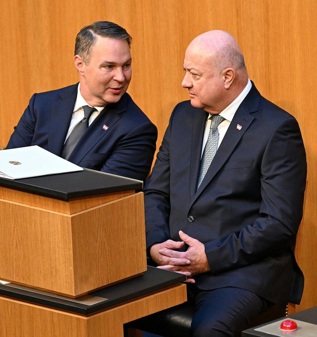 Zwei Männer in Anzügen sitzen nebeneinander und unterhalten sich.