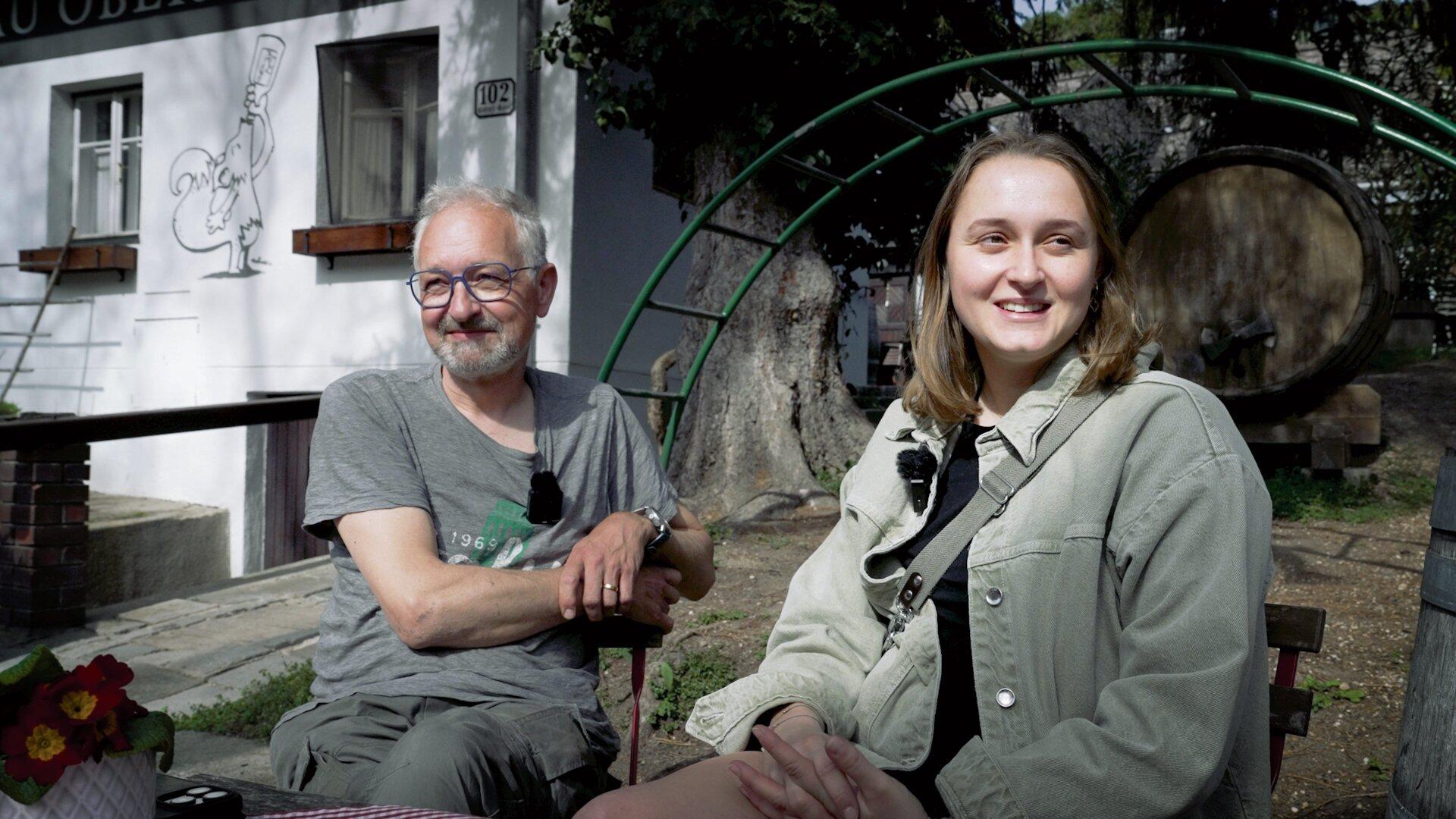 Ein Mann und eine junge Frau sitzen draußen vor einem Gebäude mit einem Fass im Hintergrund.