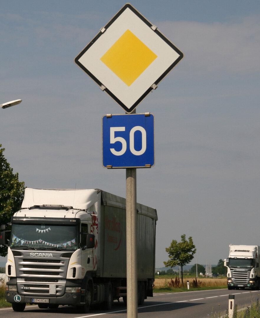 Straßenschild mit Vorfahrt und Tempo 50, daneben zwei Lastwagen auf einer Landstraße.