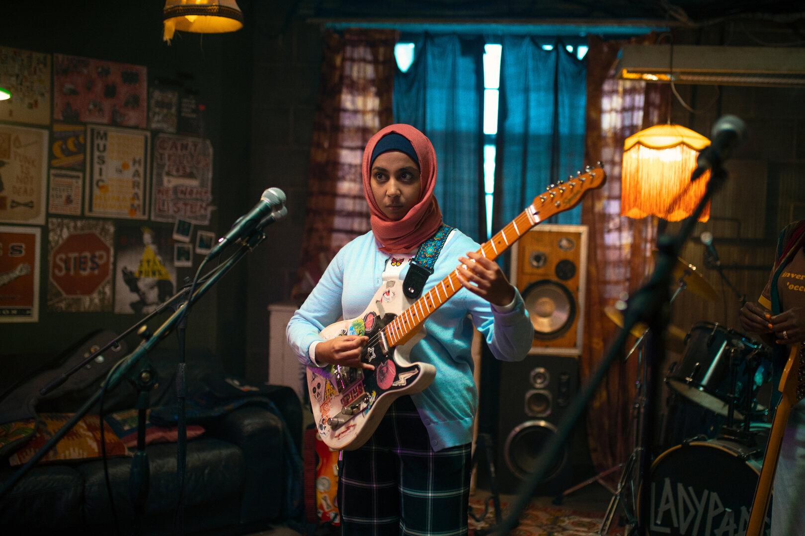 Eine Szene aus "We Are Lady Parts": Eine junge Frau (Anjana Vasan) mit Kopftuch spielt E-Gitarre in einem gemütlichen, mit Postern dekorierten Proberaum.