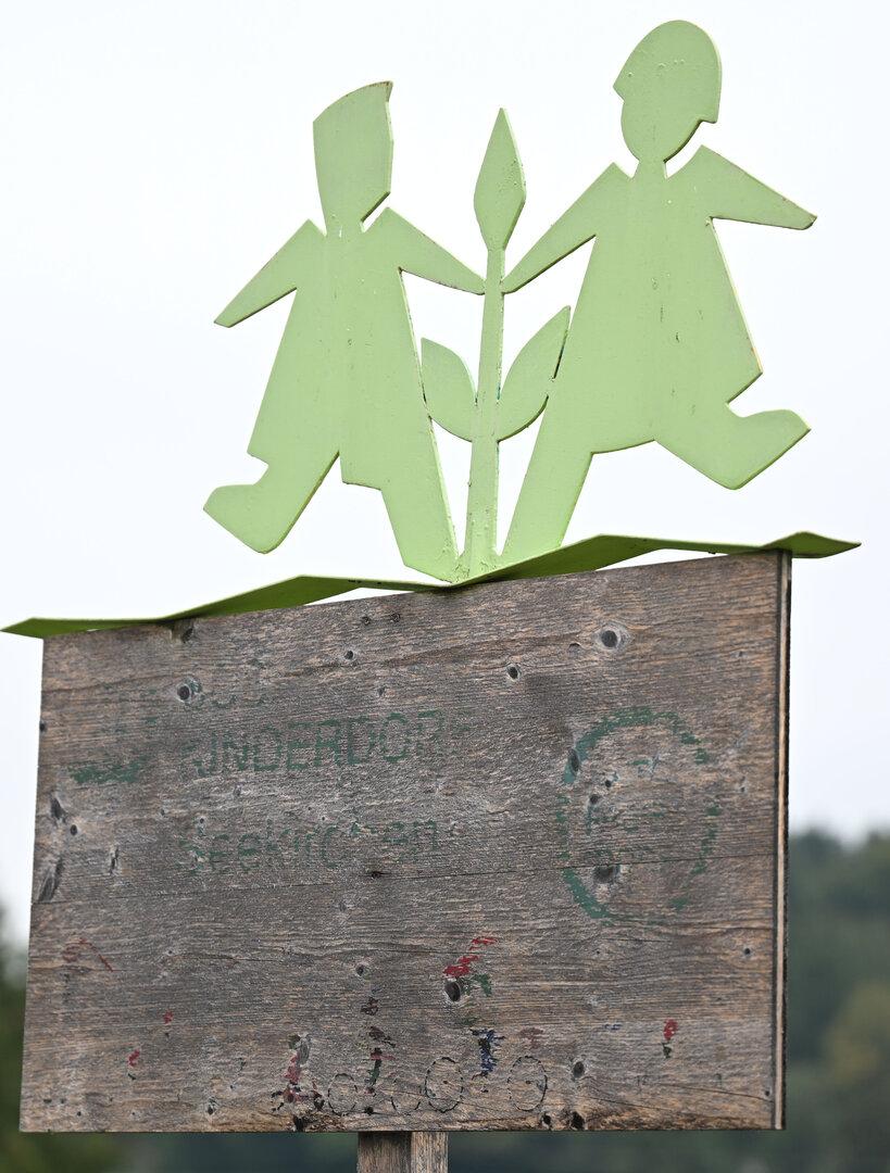 Zwei stilisierte grüne Kinderfiguren halten eine Pflanze auf einem verwitterten Holzschild.