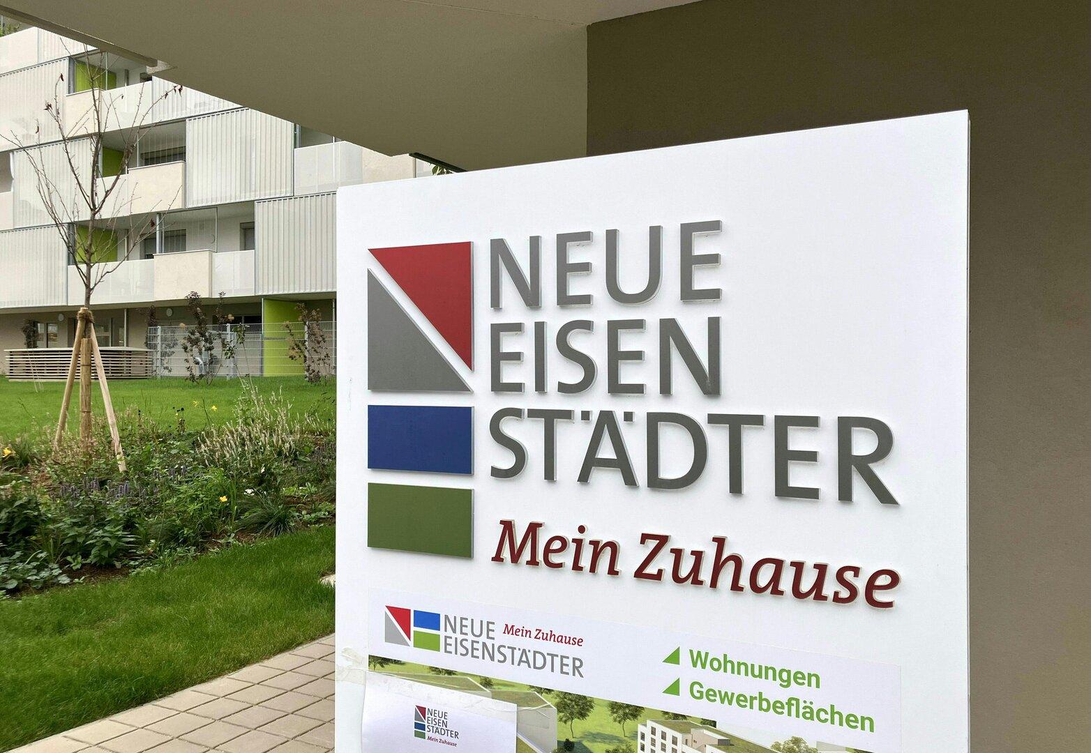 Ein Schild wirbt für "Neue Eisenstädter - Mein Zuhause" vor einem Wohngebäude.