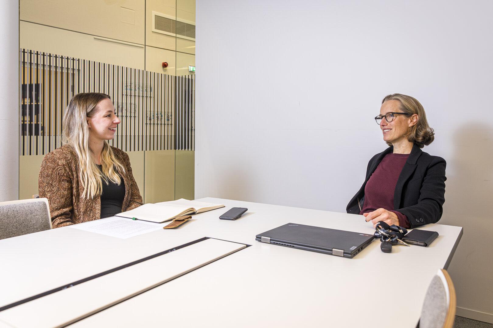 Zwei Frauen sitzen sich an einem Tisch gegenüber und unterhalten sich freundlich in einem modernen Büro.