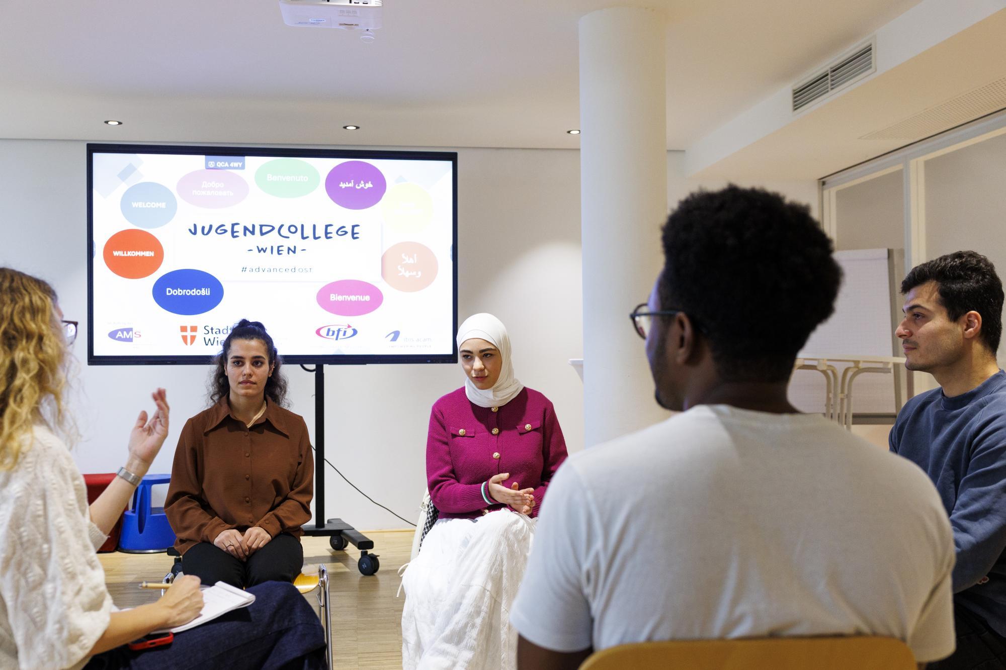 Eine Gruppe von Personen sitzt in einem Raum vor einer Leinwand des Jugendcollege Wien.