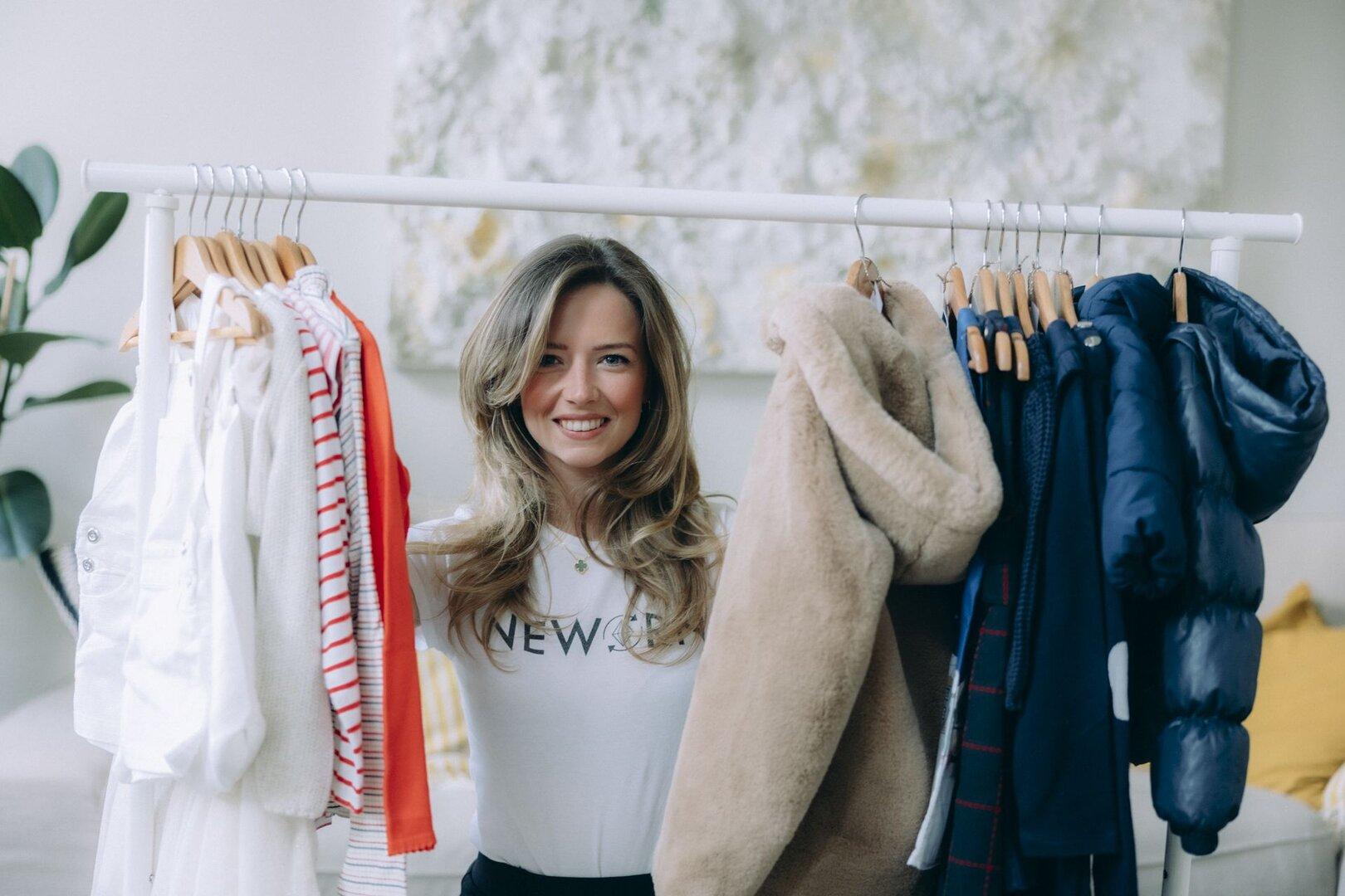 Neworn-Gründerin Caroline Schober Eine lächelnde Frau steht hinter einer Kleiderstange mit verschiedenen hellen und dunklen Kleidungsstücken.