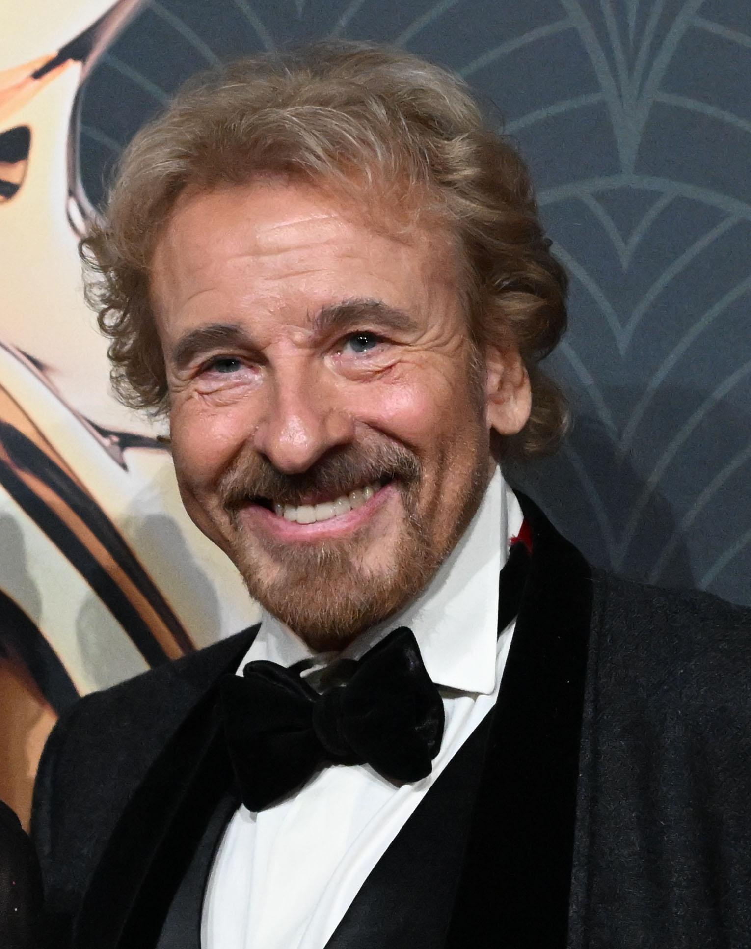 Nahaufnahme von Thomas Gottschalk mit Fliege und dunklem Jackett vor gemustertem Hintergrund.