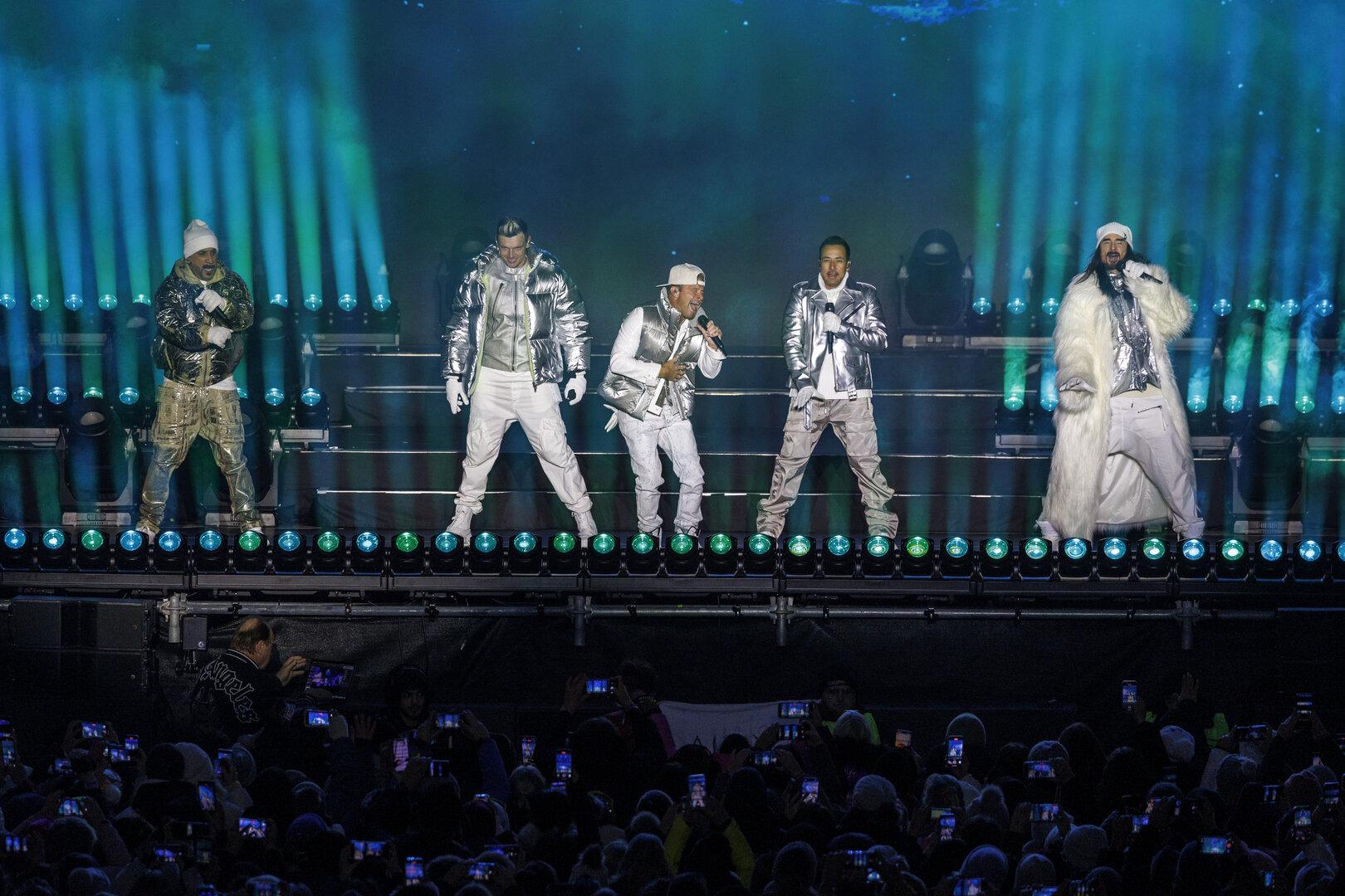 SKI-OPENING: KONZERT BACKSTREET BOYS: MCLEAN / CARTER / LITTRELL / DOROUGH / RICHARDSON Fünf Sänger in silbernen und weißen Outfits treten auf einer Bühne mit bunten Lichtern vor großem Publikum auf.