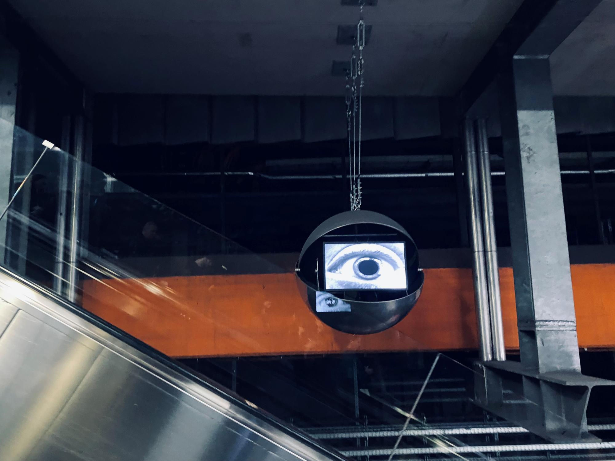 Ein Bildschirm mit einem großen, schwarz-weißen Auge hängt an Ketten in einer U-Bahn-Station.
