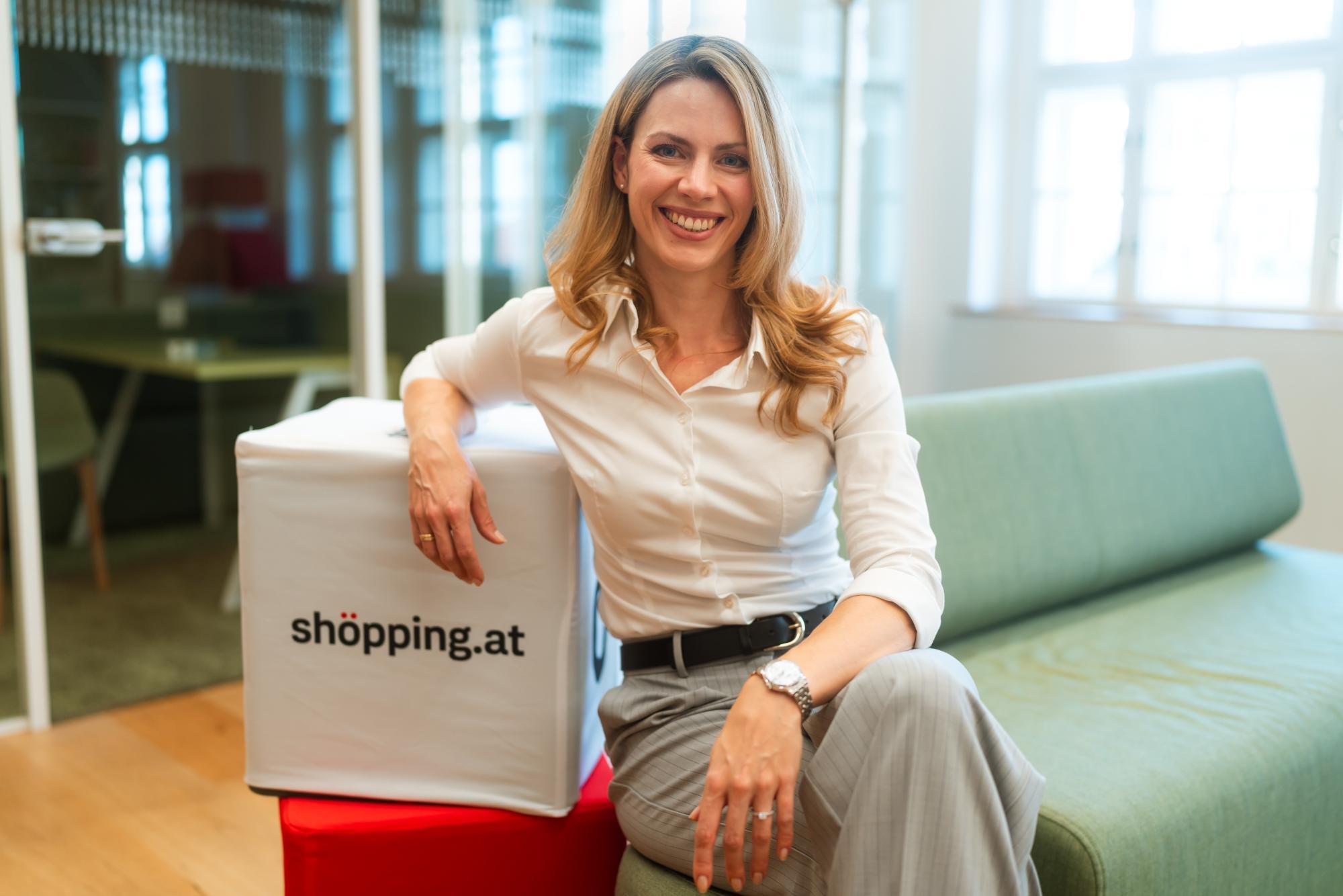 Eine Frau sitzt lächelnd auf einem Sofa und lehnt an einem Würfel mit der Aufschrift "shöpping.at"