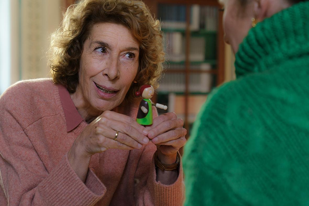 Eine ältere Frau im rosa Pullover hält eine kleine Holzpuppe in den Händen.