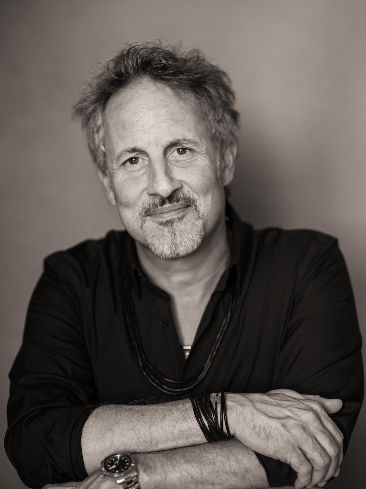 Ein Mann mit grauem Haar und Bart trägt ein dunkles Hemd und mehrere Armbänder, er blickt freundlich in die Kamera.