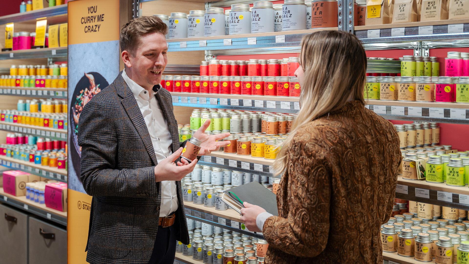 Zwei Personen unterhalten sich vor einem Regal mit bunten Gewürzdosen, eine Person hält eine Dose in der Hand.