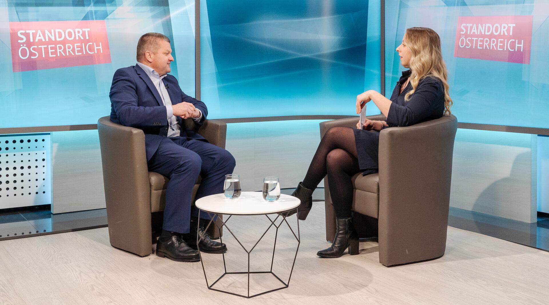 Zwei Personen sitzen in Sesseln vor dem Hintergrund "Standort Österreich" und unterhalten sich.