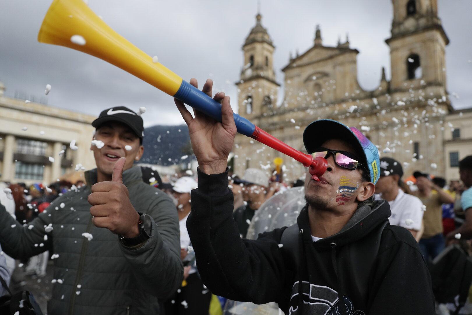 Zwei Männer feiern mit einer bunten Tröte und Daumen hoch vor einer historischen Kirche, umgeben von Konfetti und Menschenmenge.