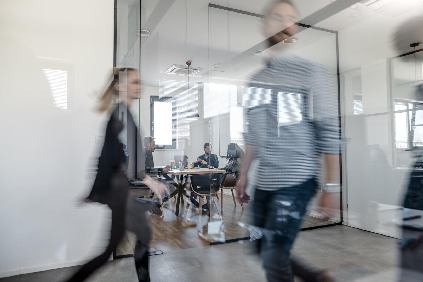 Im Büro gehen verschwommene Personen vorbei, während andere an einem Tisch sitzen.