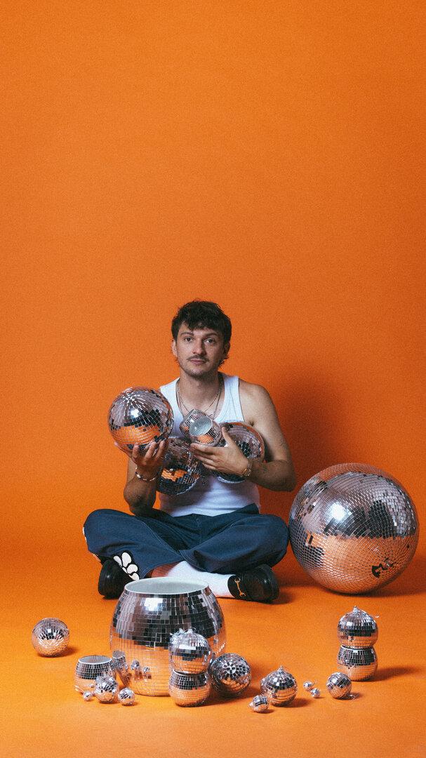 Der Musiker Rian sitzt vor orangefarbenem Hintergrund und hält mehrere Discokugeln verschiedener Größen in den Händen und auf dem Boden.