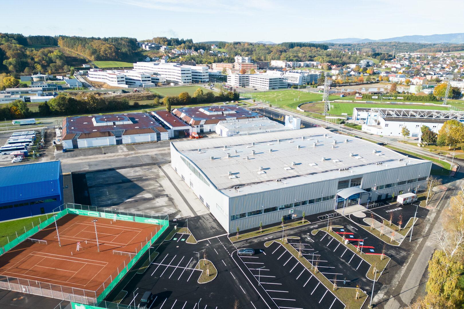 Luftaufnahme eines Gewerbegebiets mit Hallen, Tennisplätzen, Parkplätzen, Sportfeldern und Wohnhäusern im Hintergrund.