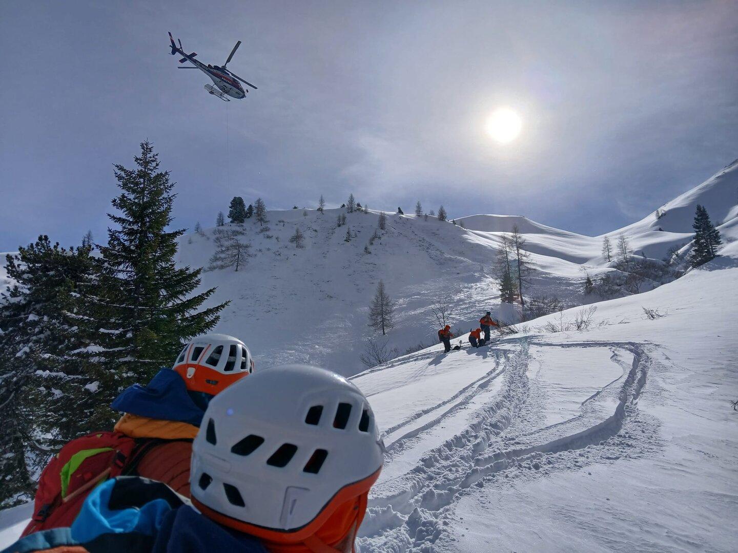 Ein Hubschrauber fliegt über eine verschneite Berglandschaft, während Bergretter zu sehen sind.