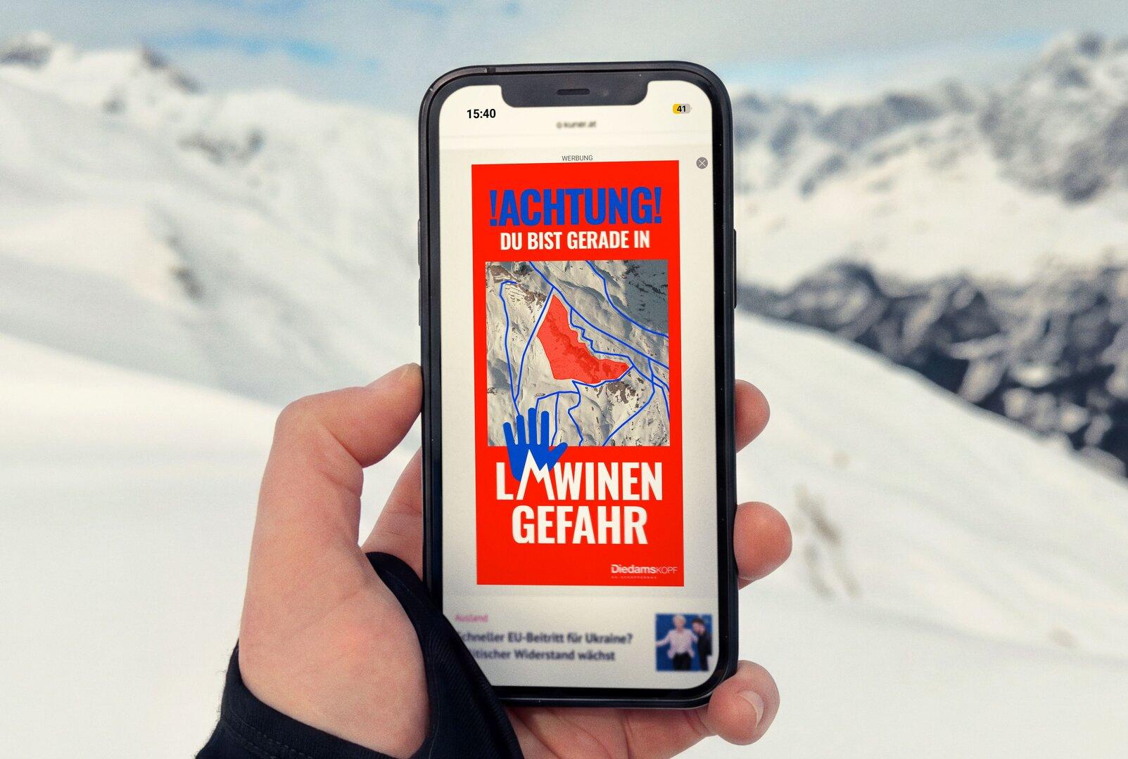 Eine Hand hält ein Smartphone, das eine Lawinengefahr-Warnung für Diedamskopf anzeigt.