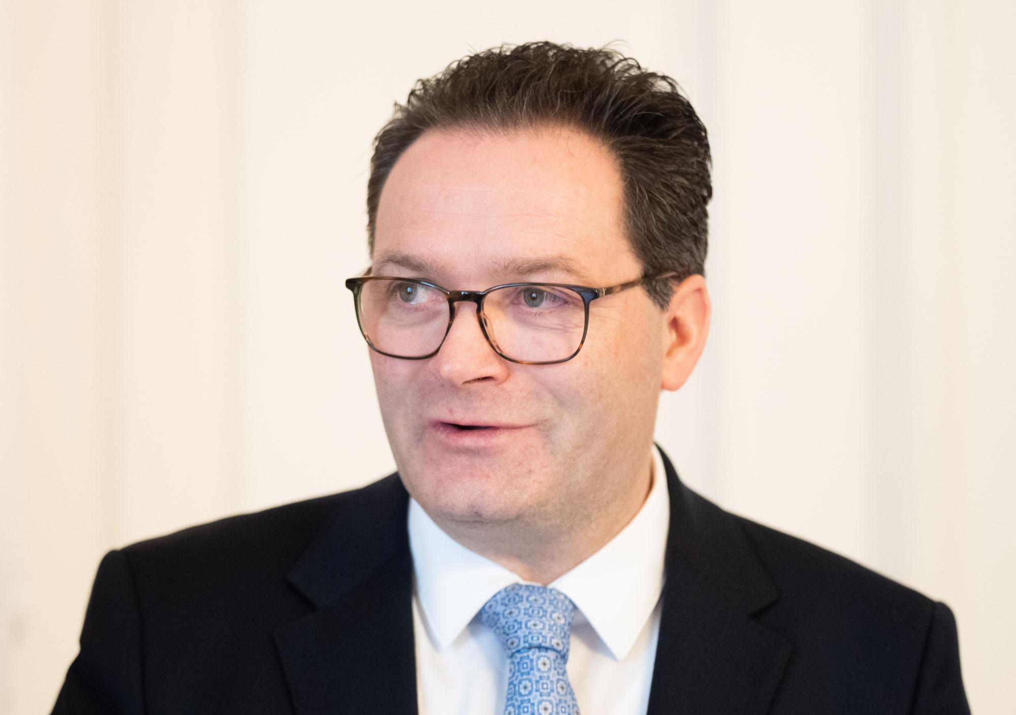 Mann mit dunklem Anzug, weißem Hemd, gemusterter Krawatte und Brille vor hellem Hintergrund.