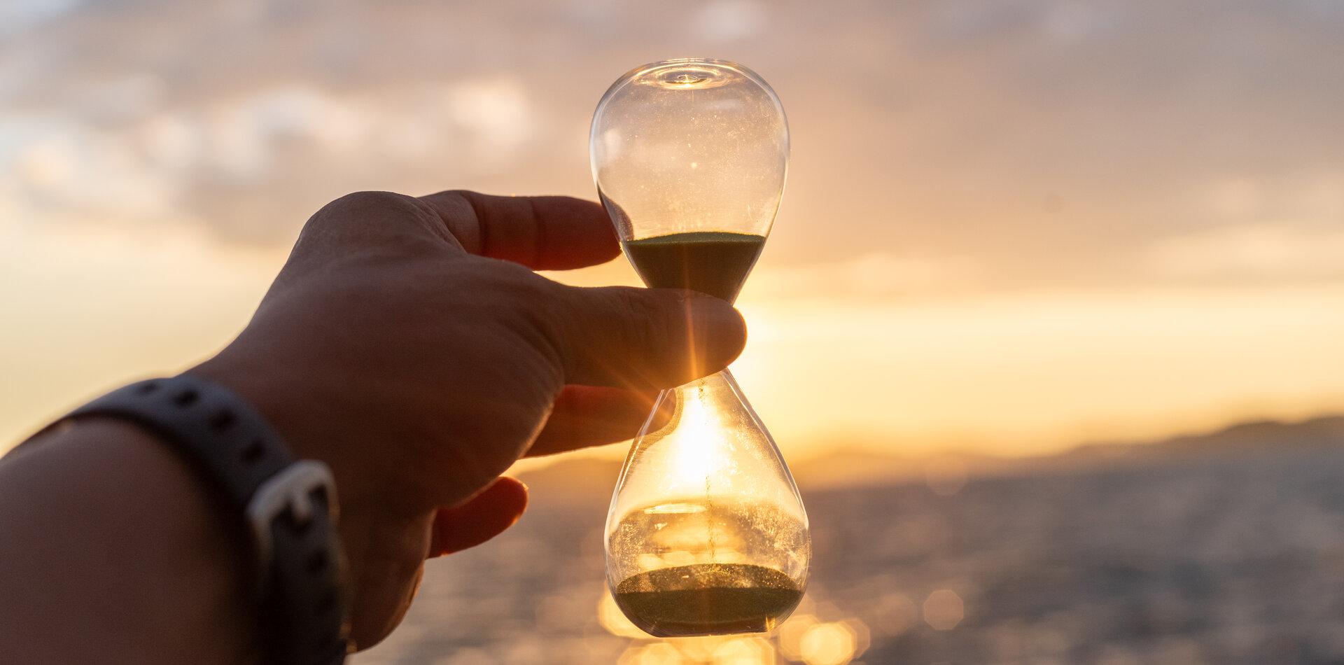 Eine Hand hält eine Sanduhr vor einem sonnigen Himmel.
