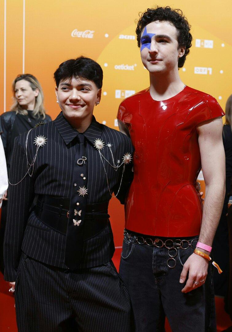 Zwei Personen posieren vor orangefarbenem Hintergrund, eine trägt einen Nadelstreifenanzug mit Schmuck, die andere ein rotes Oberteil und Gesichtsbemalung.