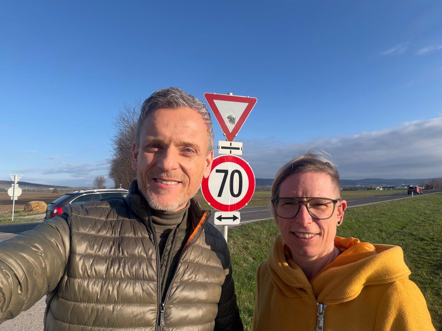 Zwei Personen stehen bei Sonnenschein neben einer Landstraße vor einem Vorfahrt-achten- und einem 70-km/h-Schild.