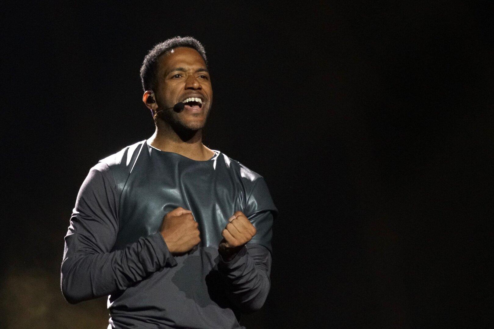 Cesár Sampson mit Mikrofon am Ohr singt mit geballten Fäusten vor dunklem Hintergrund.