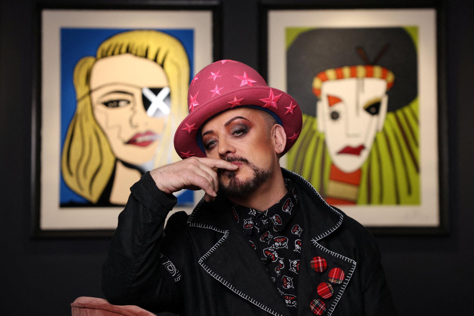 Boy George posiert mit Sternen-Hut vor zwei gerahmten Kunstwerken.