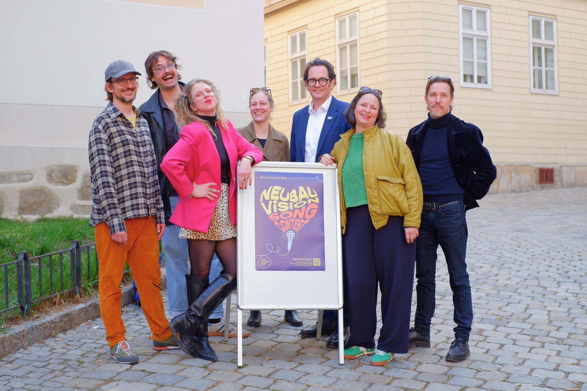 Mehrere bunt gekleidete Menschen, darunter Neubau Bezirksvorsteher Reiter, stehen vor einem Plakatschild auf Pflastersteinen