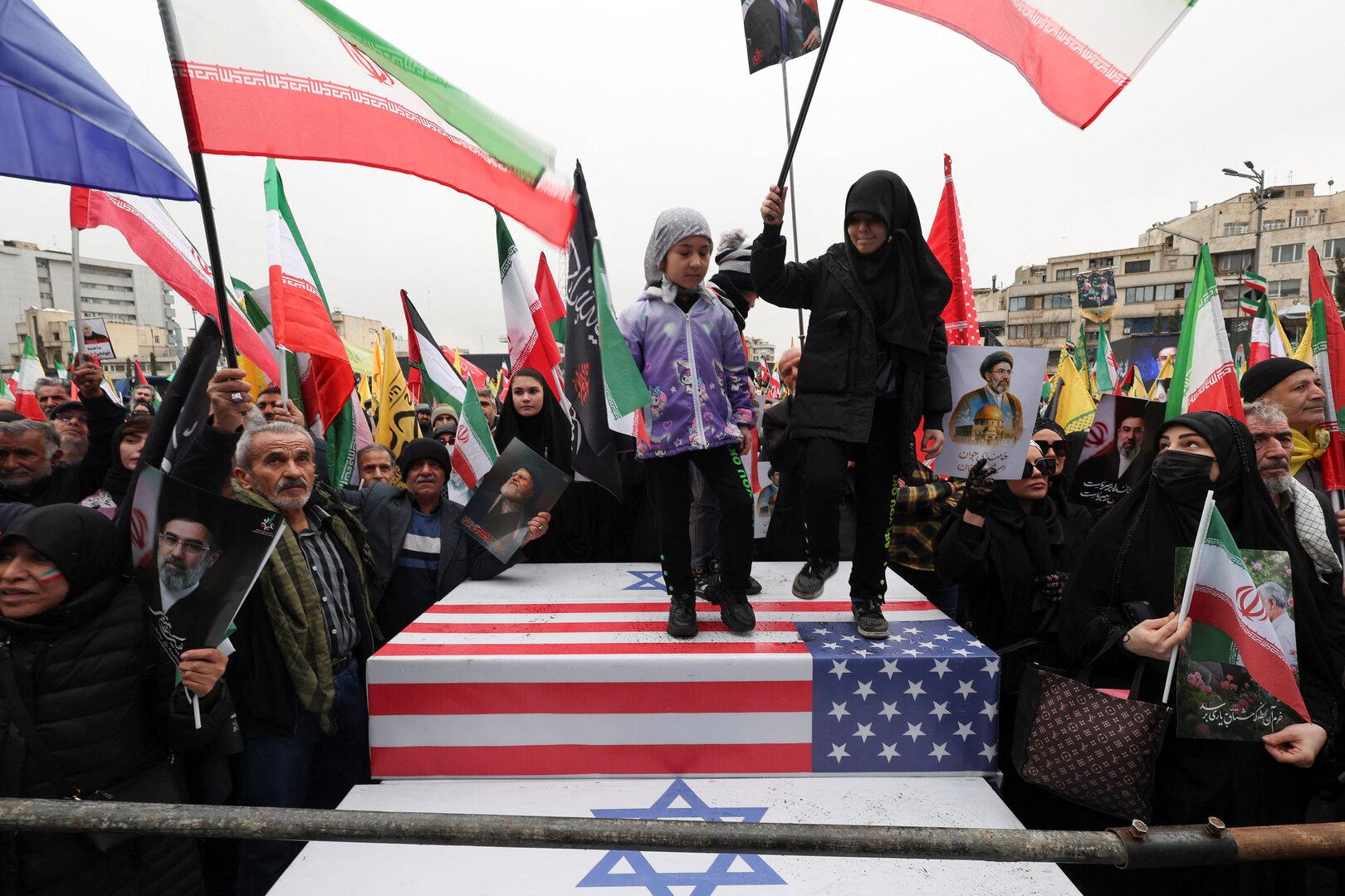 Zwei Kinder stehen auf einer Plattform mit US-Flagge und Davidstern, umgeben von Menschen mit iranischen Fahnen und Porträts.