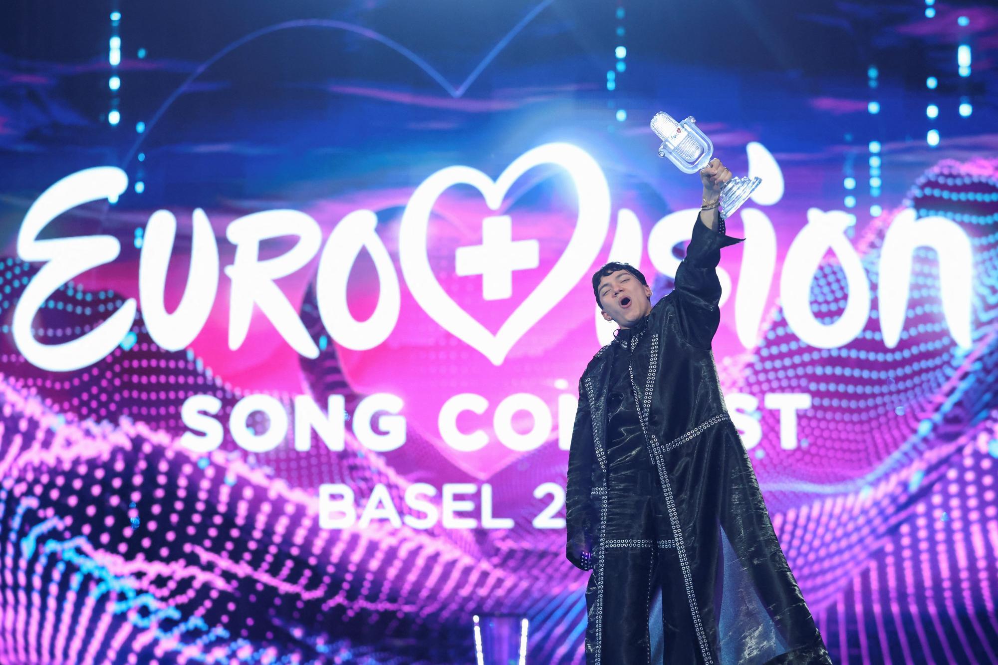 Eine Person hält jubelnd einen gläsernen Mikrofon-Pokal vor einer bunten Eurovision Song Contest Bühne hoch.