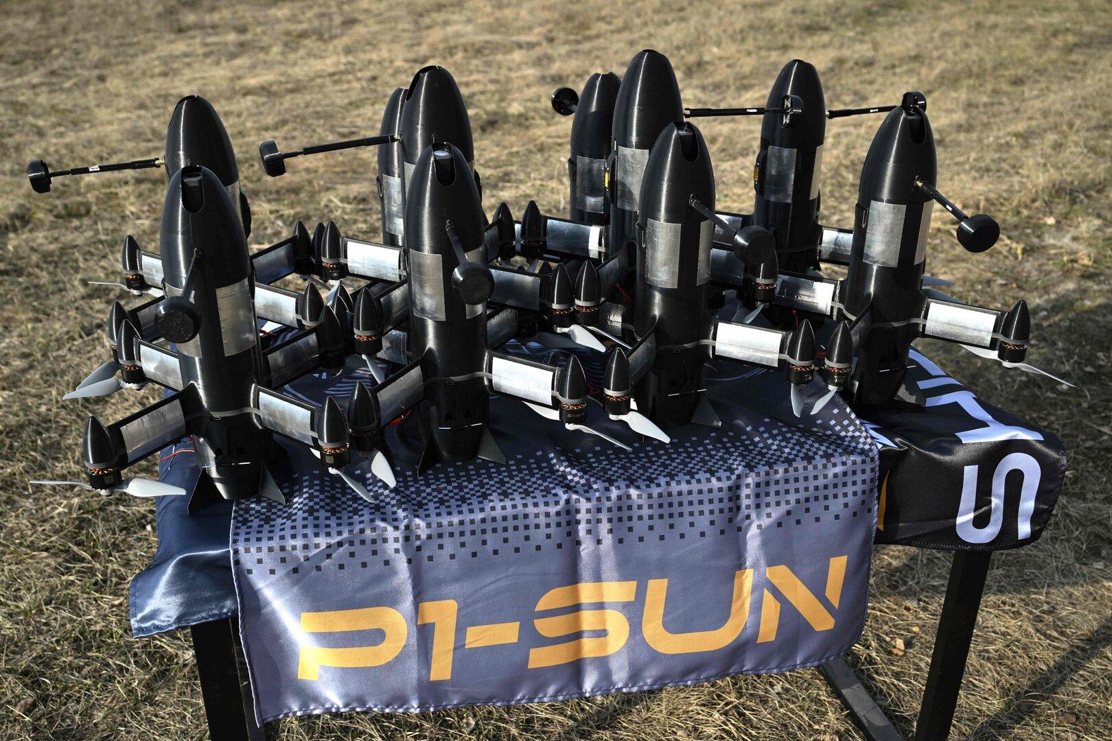 Mehrere schwarze Drohnen mit Propellern stehen auf einem Tisch im Freien auf einer blauen Decke mit gelber Aufschrift.