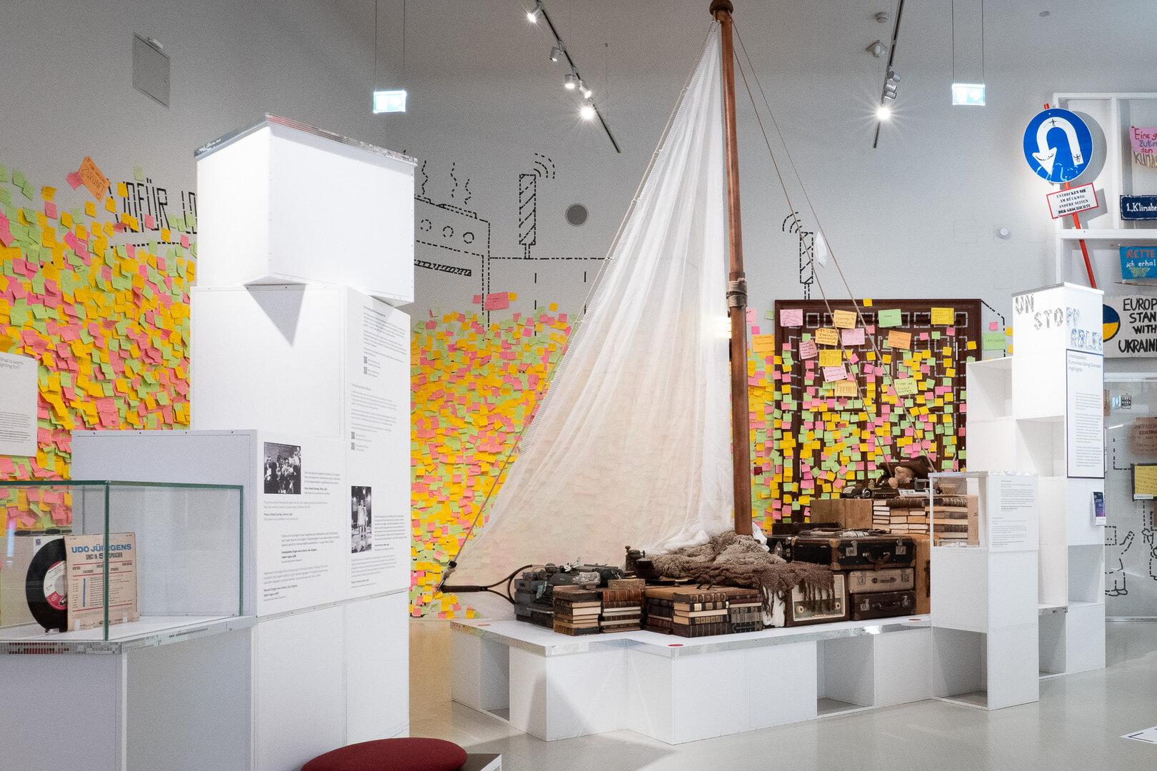 Ein Segelboot mit Büchern, Koffern und Alltagsgegenständen steht in einem Ausstellungsraum, die Wände sind mit bunten Notizzetteln bedeckt.