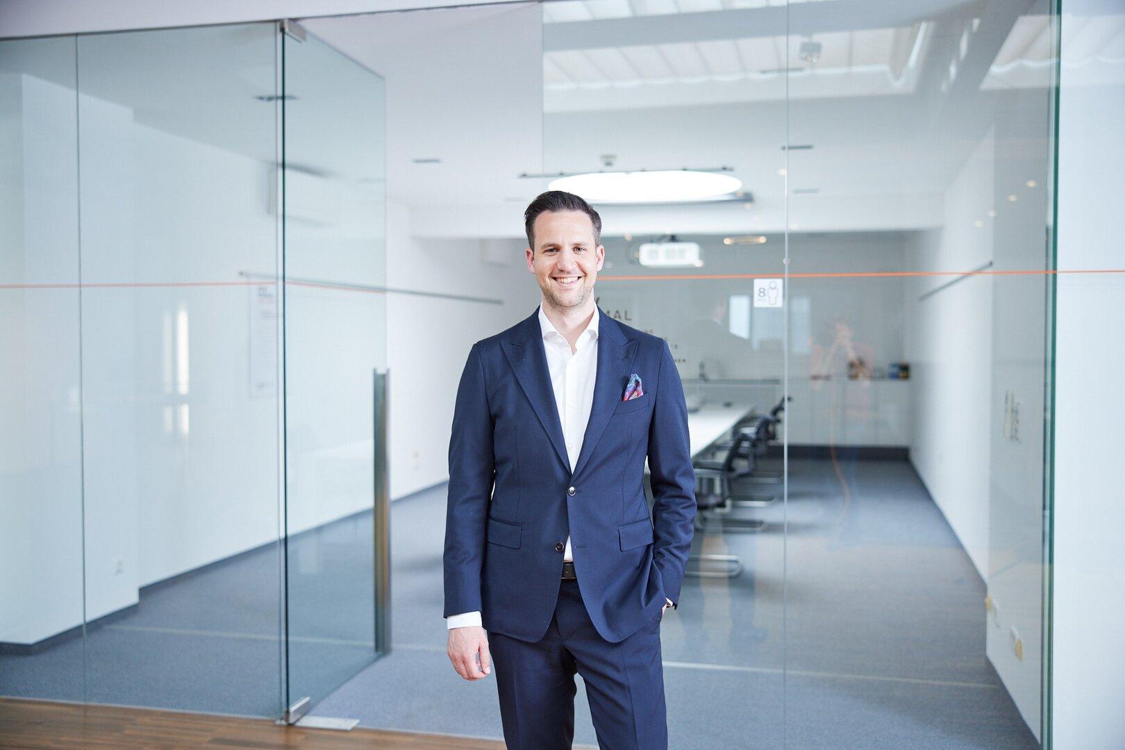 Ein Mann im Anzug steht lächelnd in einem modernen, hellen Büro mit Glastüren und Konferenztisch.