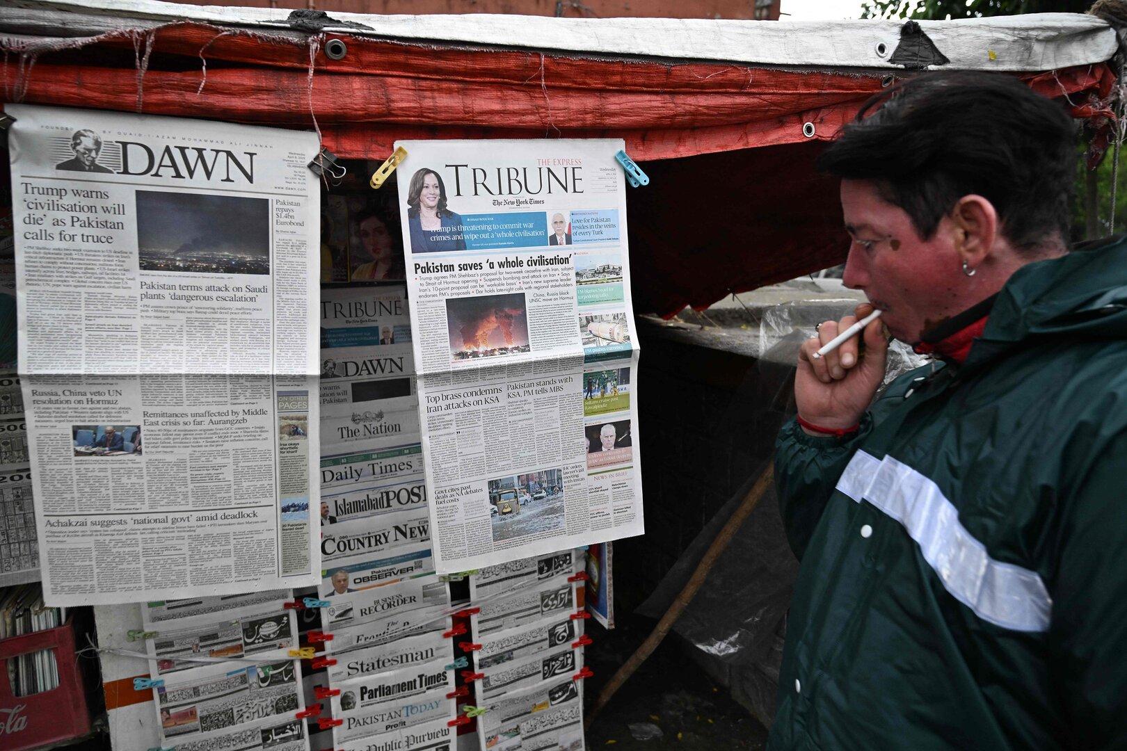 Eine Person mit Zigarette betrachtet eine Auswahl an Zeitungen, die an einem Straßenstand aufgehängt sind.