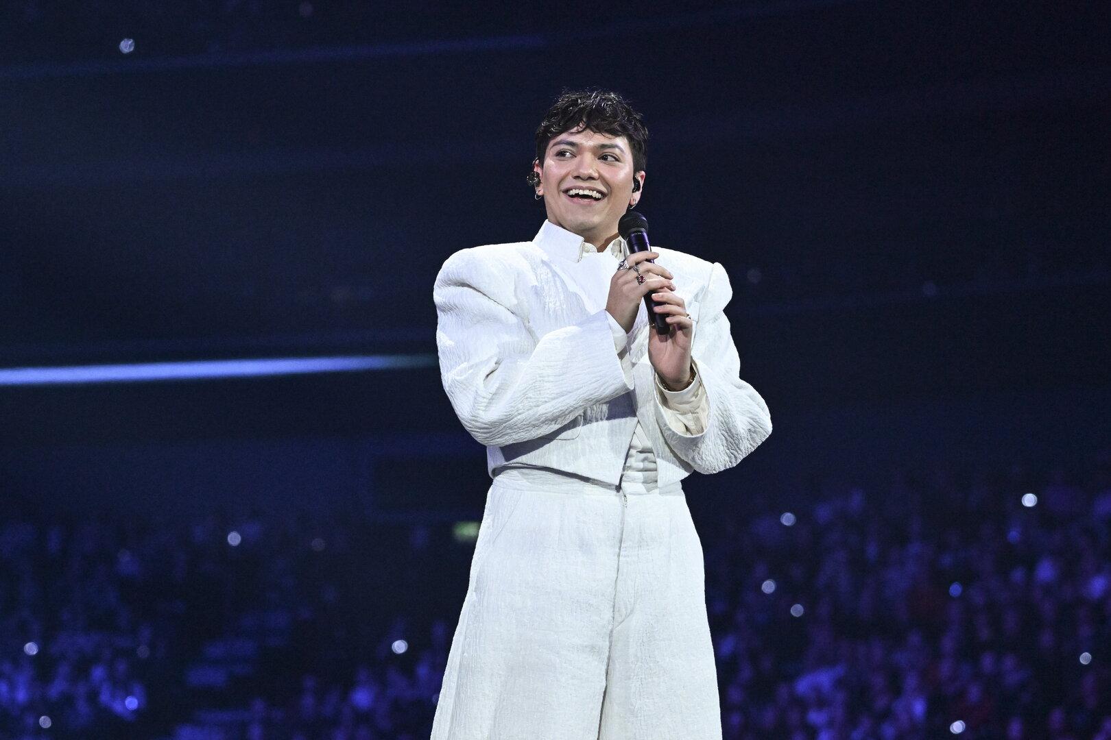 Eine Person in weißem Anzug hält ein Mikrofon und lächelt auf einer Bühne vor großem Publikum.