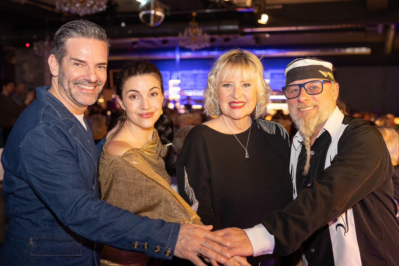 Vier lächelnde Personen legen ihre Hände übereinander in einem festlichen Rahmen.