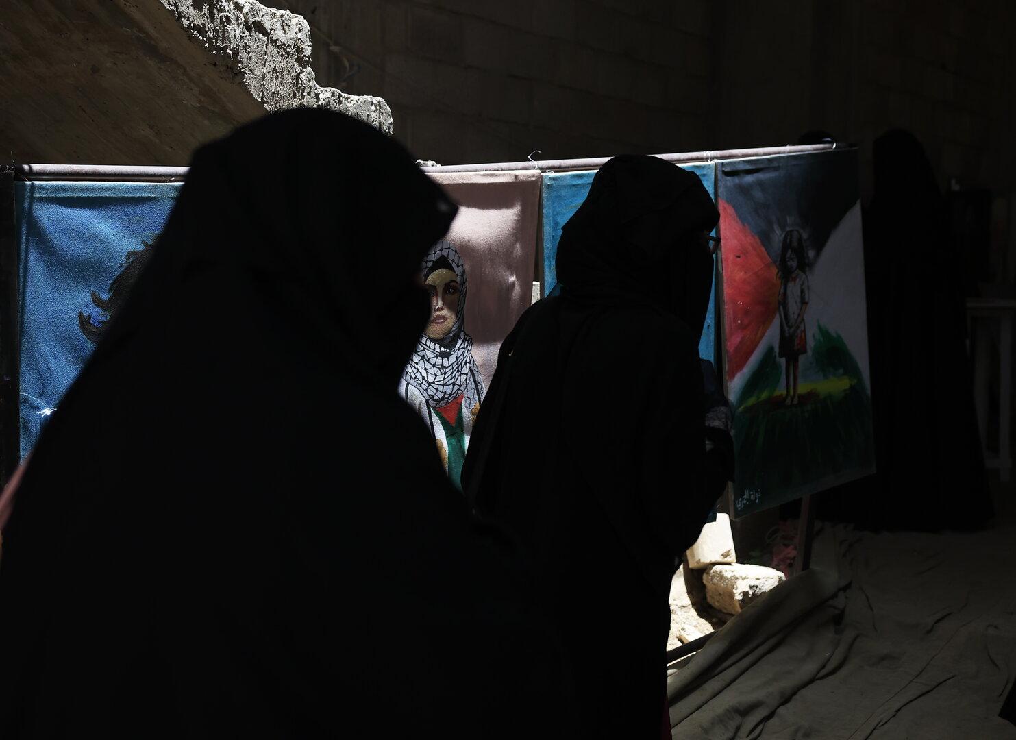 Zwei Frauen in dunkler Kleidung stehen vor Gemälden, darunter Porträts und ein Kind vor einer Flagge.