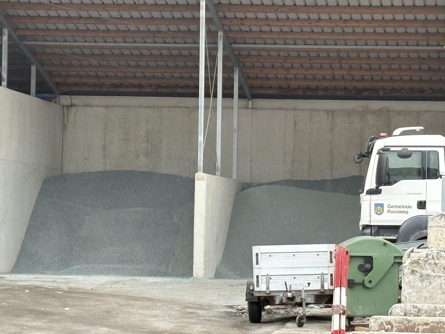 Zwei Haufen Streusalz oder Splitt unter einem Dach, daneben ein Kommunalfahrzeug mit Anhänger und Müllcontainer.